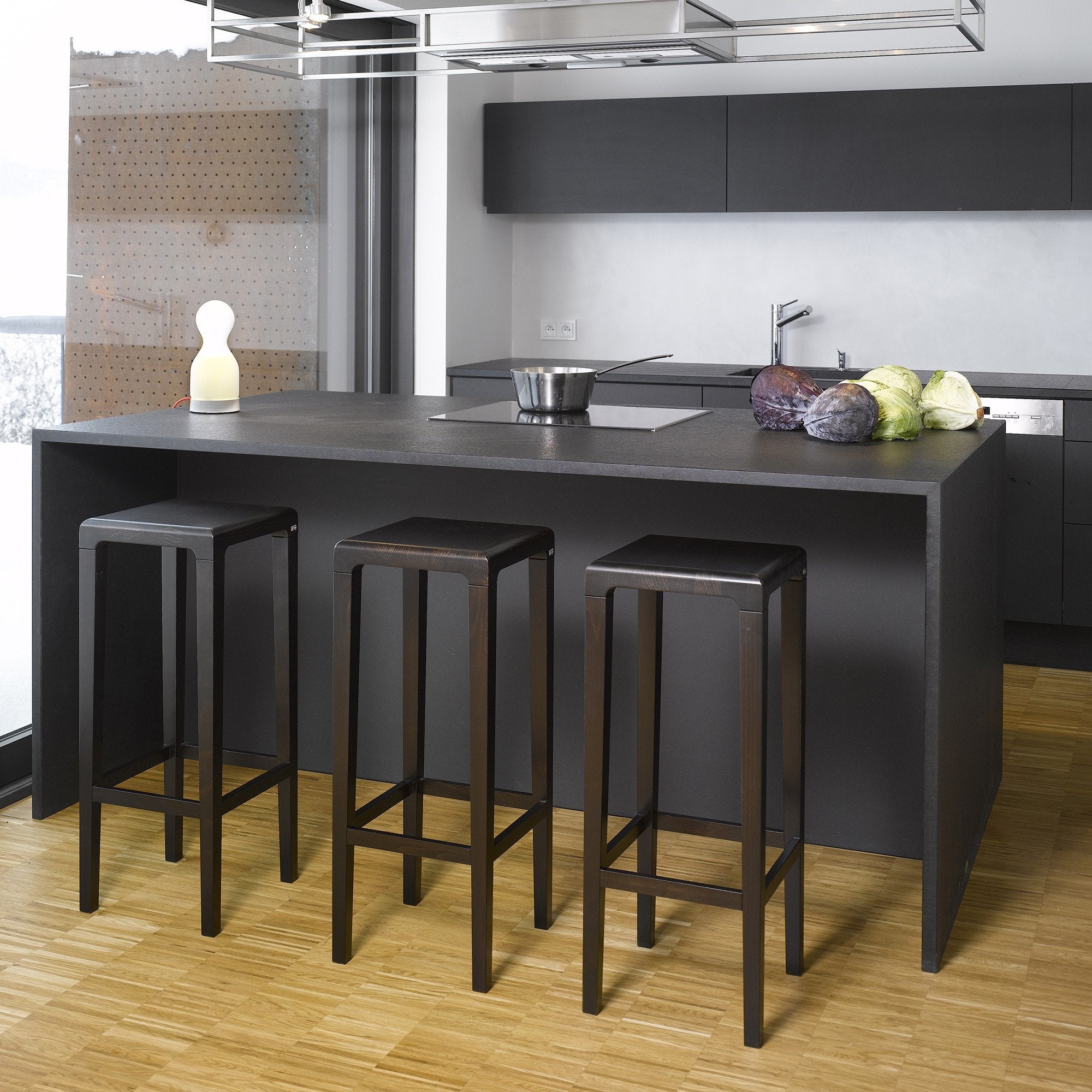 A modern kitchen showcases a sleek black island surrounded by three Barstool Rioja from TON. The island is topped with a small white lamp and vegetables, while elegant black cabinets, warm beech-like wooden flooring, and a large window on the left complete the space.