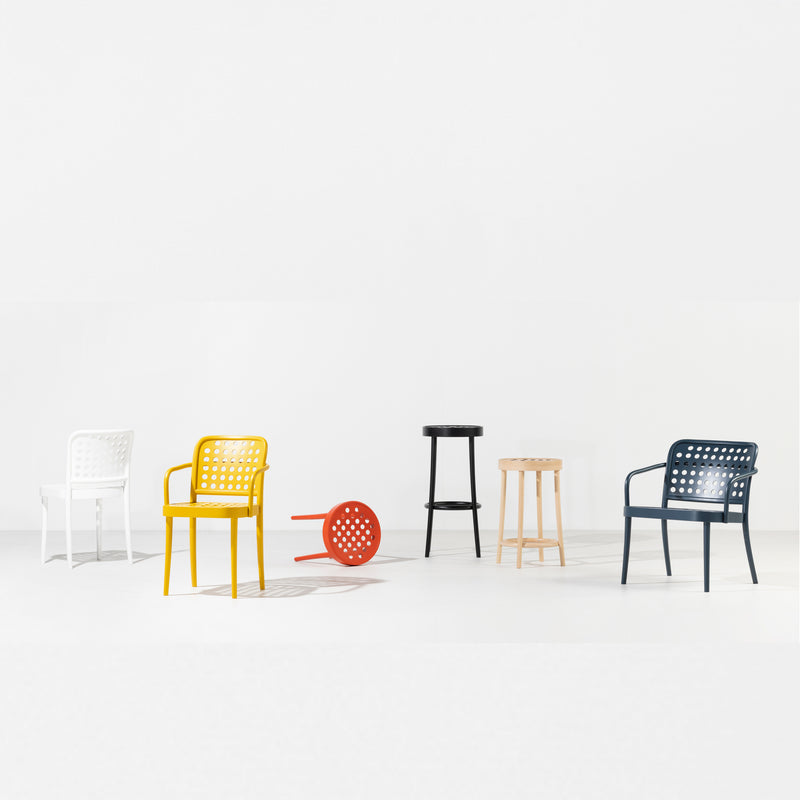 A minimalist scene captures Scandinavian minimalism: five artfully arranged chairs and stools—a white Lounge Chair 822 by TON, a yellow chair, an overturned red chair, a black stool, and a light wooden stool—on a pristine white background, showcasing the elegance of wood-bending technology by TONDesigner Studio.