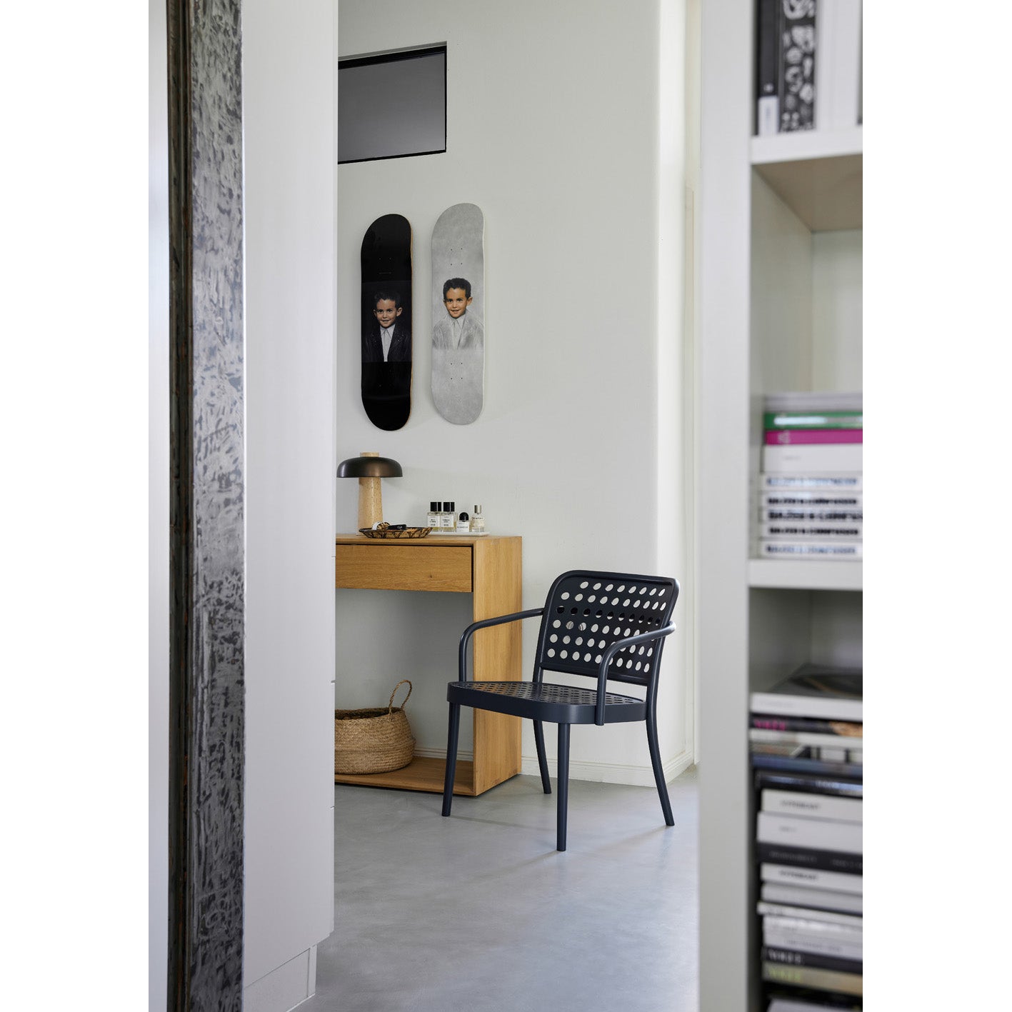 A modern interior showcases a wooden desk, a Lounge Armchair 822 by TON, and a lamp. Above the desk, two skateboard decks with portraits add character. A woven basket is placed below it. Embracing Scandinavian minimalism, a partly visible bookshelf on the right holds neatly stacked items.