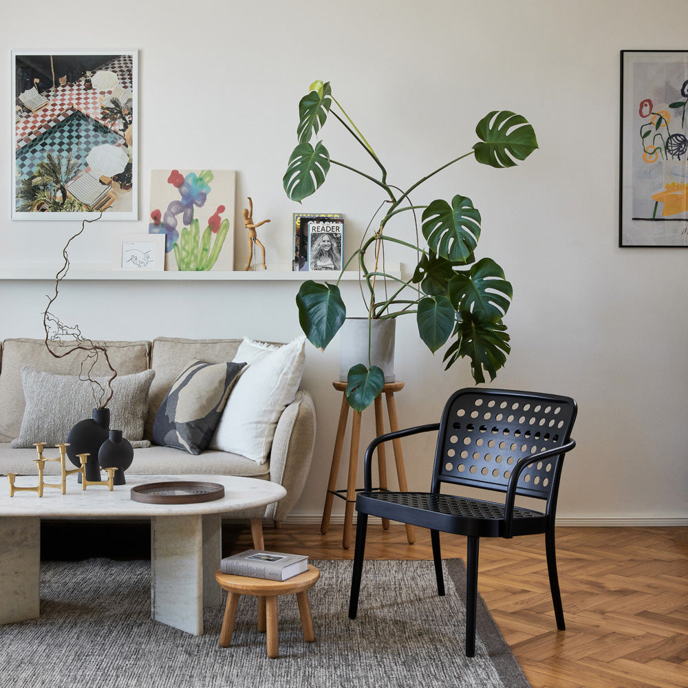 A stylish living room with a beige sofa, round coffee table, and TONs Lounge Armchair 822 in black embodies Scandinavian minimalism. A large potted monstera plant adds greenery, while art and decor adorn the walls and shelves, creating a cozy, contemporary vibe akin to Claesson Koivisto Runes designs.