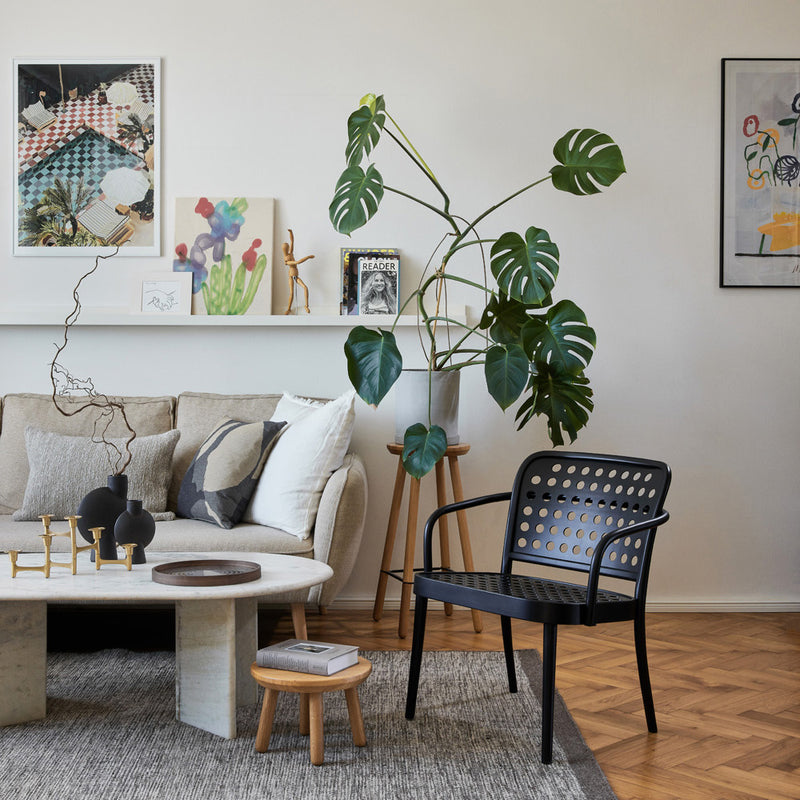 A stylish living room with a beige sofa, round coffee table, and TONs Lounge Armchair 822 in black embodies Scandinavian minimalism. A large potted monstera plant adds greenery, while art and decor adorn the walls and shelves, creating a cozy, contemporary vibe akin to Claesson Koivisto Runes designs.