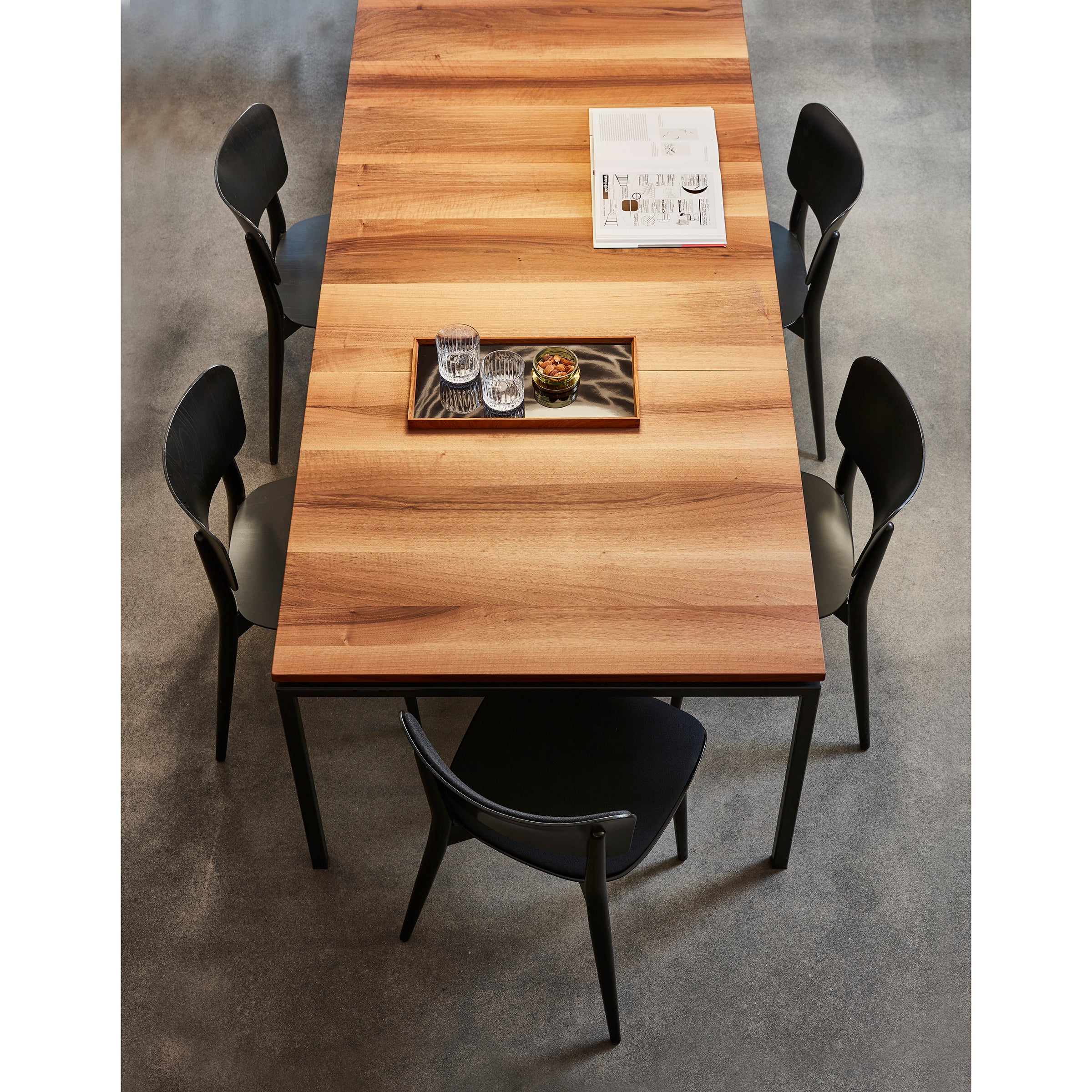 The UPW 1010 Extendable Table by WB Form, displaying superior craftsmanship, features a tray with glasses and a small plant against its gray floor setting. Surrounded by four black chairs, an open magazine lies at one end, inviting relaxation.