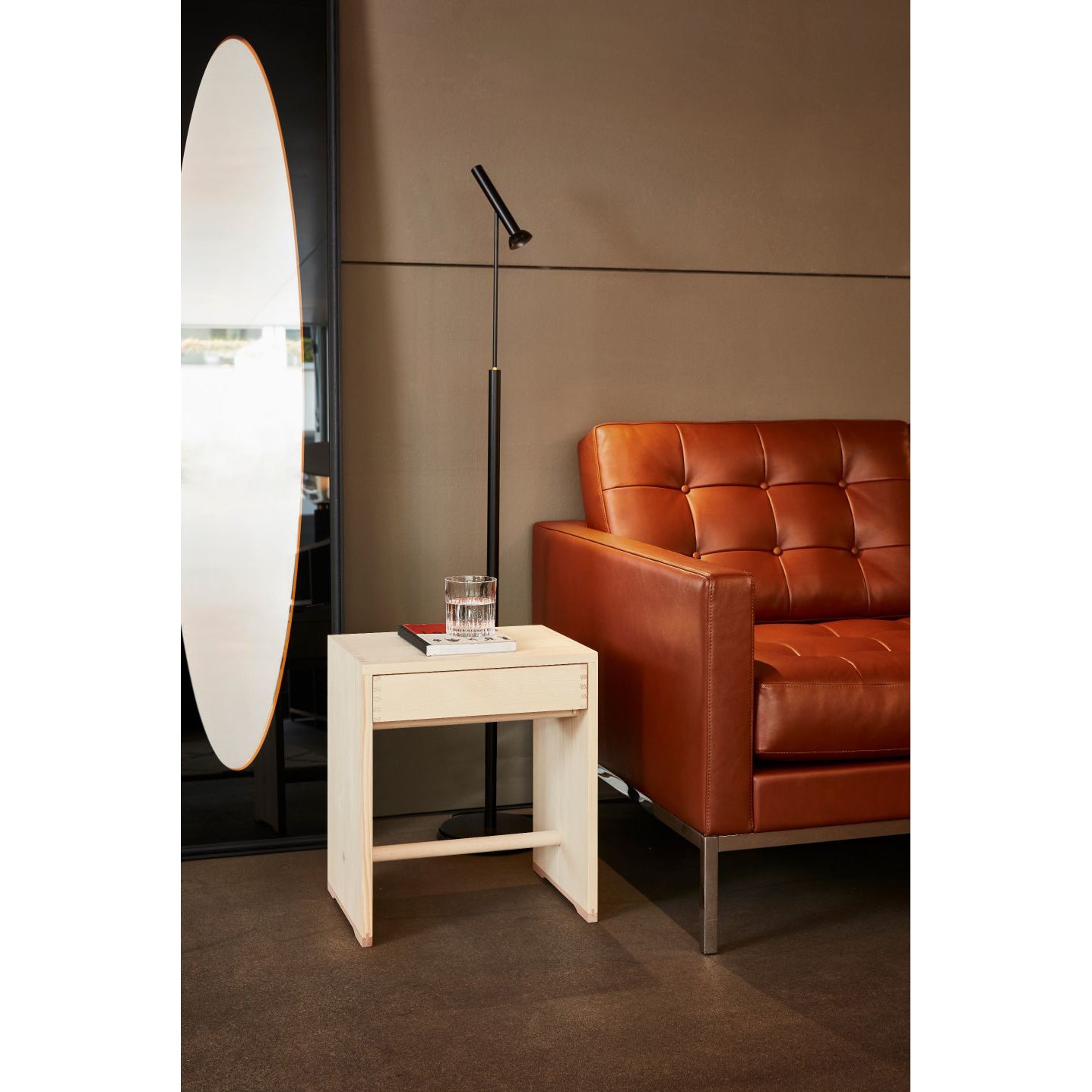 In a modern living room, a brown leather sofa sits beside a sleek white side table holding a glass of water and a closed book. Nearby stands a minimalist black floor lamp, while an Ulmer Stool with Drawer by WB Form complements the large oval mirror leaning against the dark wall.