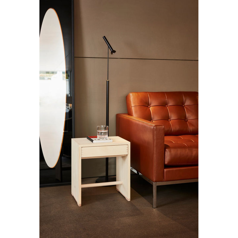 In a modern living room, a brown leather sofa sits beside a sleek white side table holding a glass of water and a closed book. Nearby stands a minimalist black floor lamp, while an Ulmer Stool with Drawer by WB Form complements the large oval mirror leaning against the dark wall.
